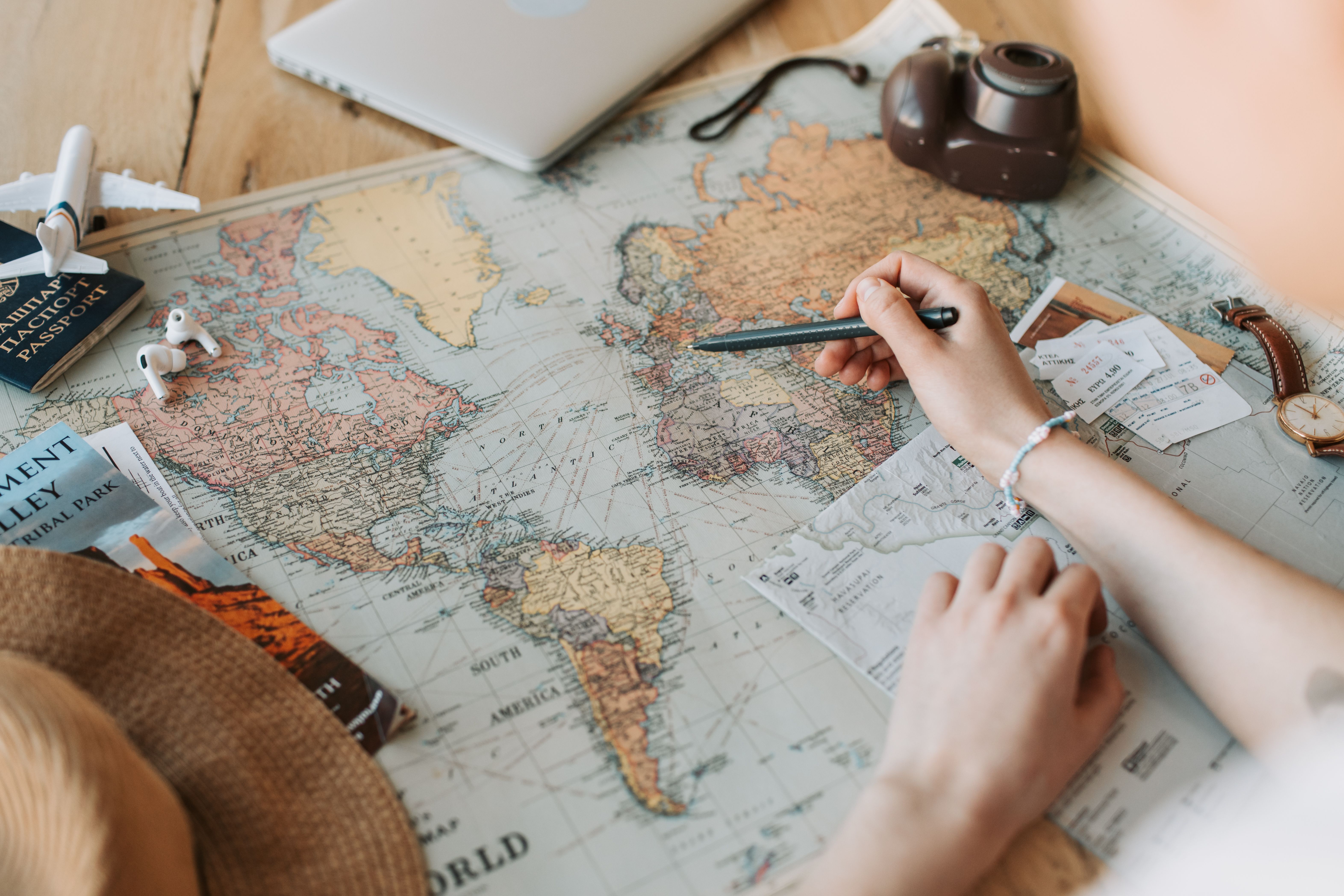 Hands marking destinations on a world map surrounded by travel essentials like a passport and camera